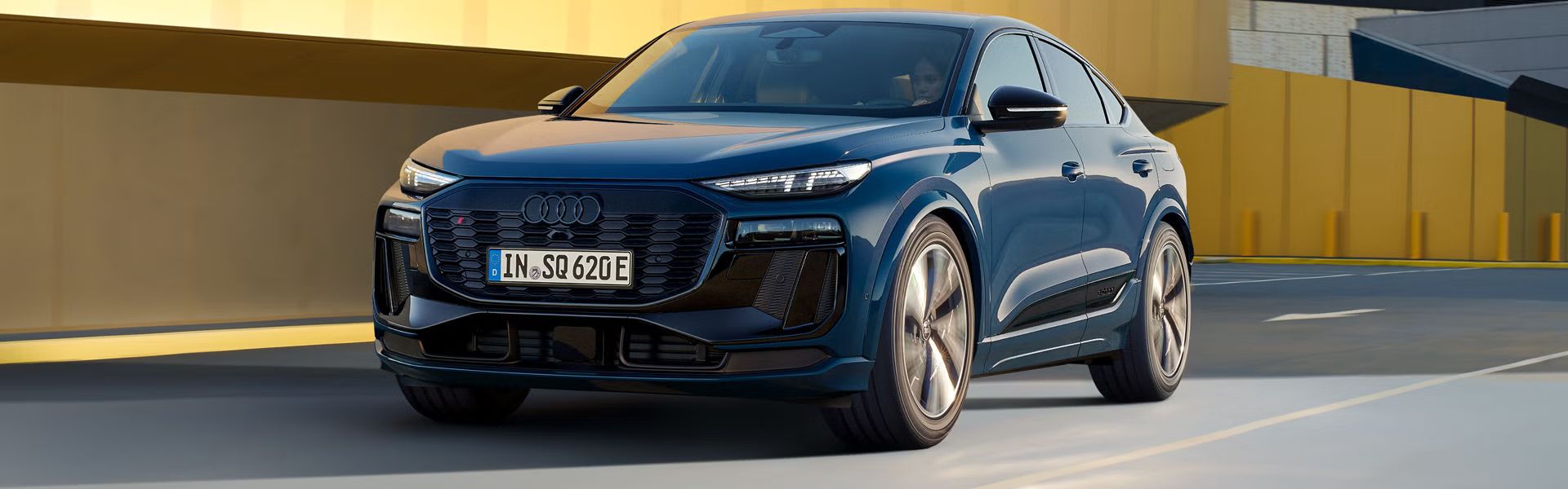 Ein blauer Audi SQ6 Sportback e-tron fährt auf einer Straße vor einem gelben Gebäude.