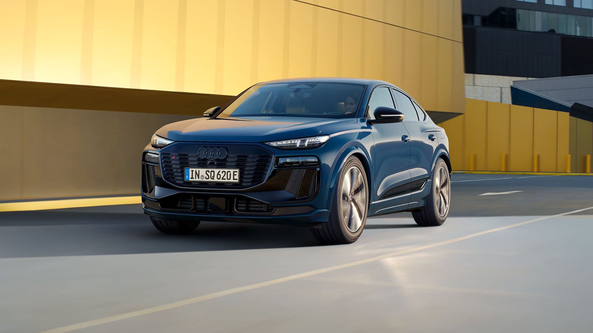 Ein blauer Audi SQ6 Sportback e-tron fährt auf einer Straße vor einem gelben Gebäude.