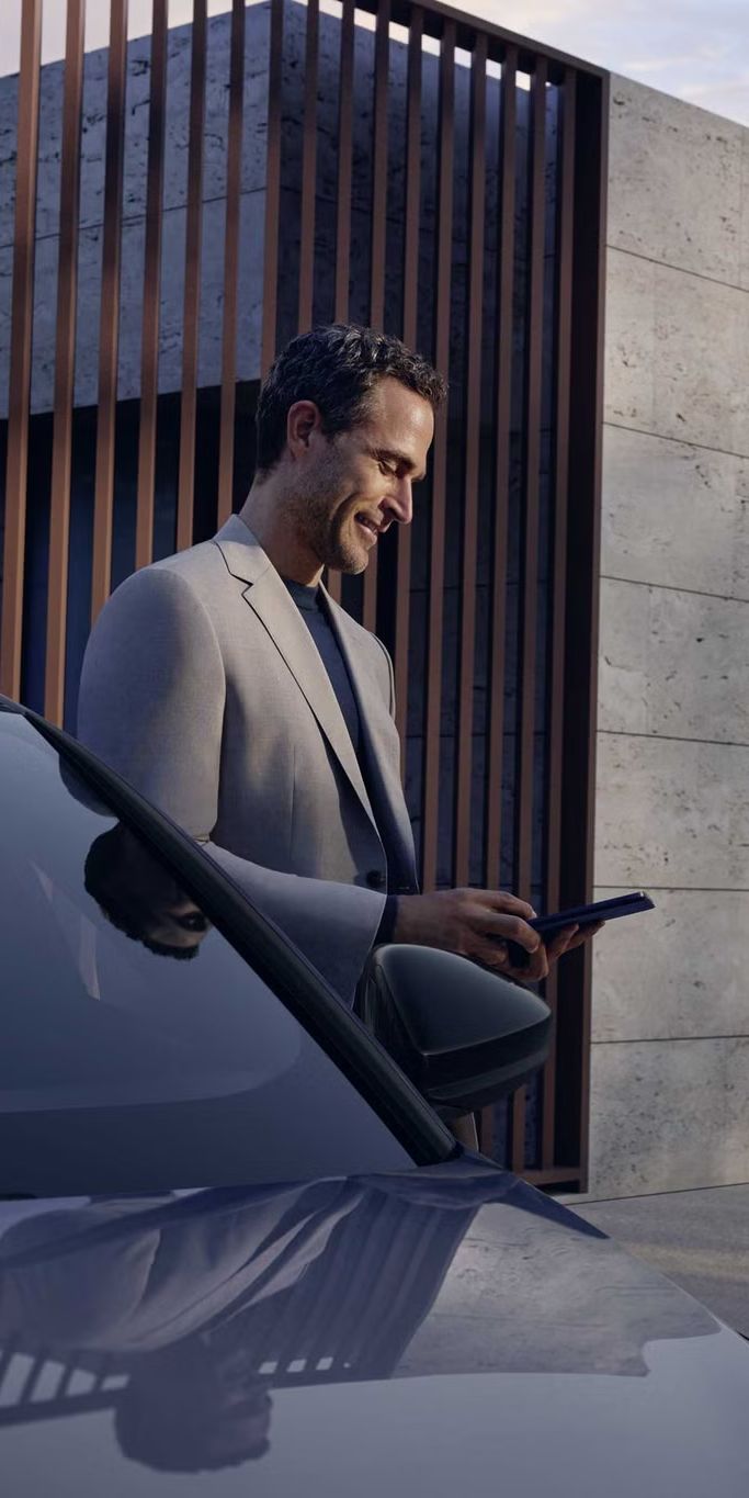 Mann mit Smartphone vor Audi, nutzt Fahrzeug-App; im Hintergrund modernes Holzgebäude und grüne Rasenfläche.