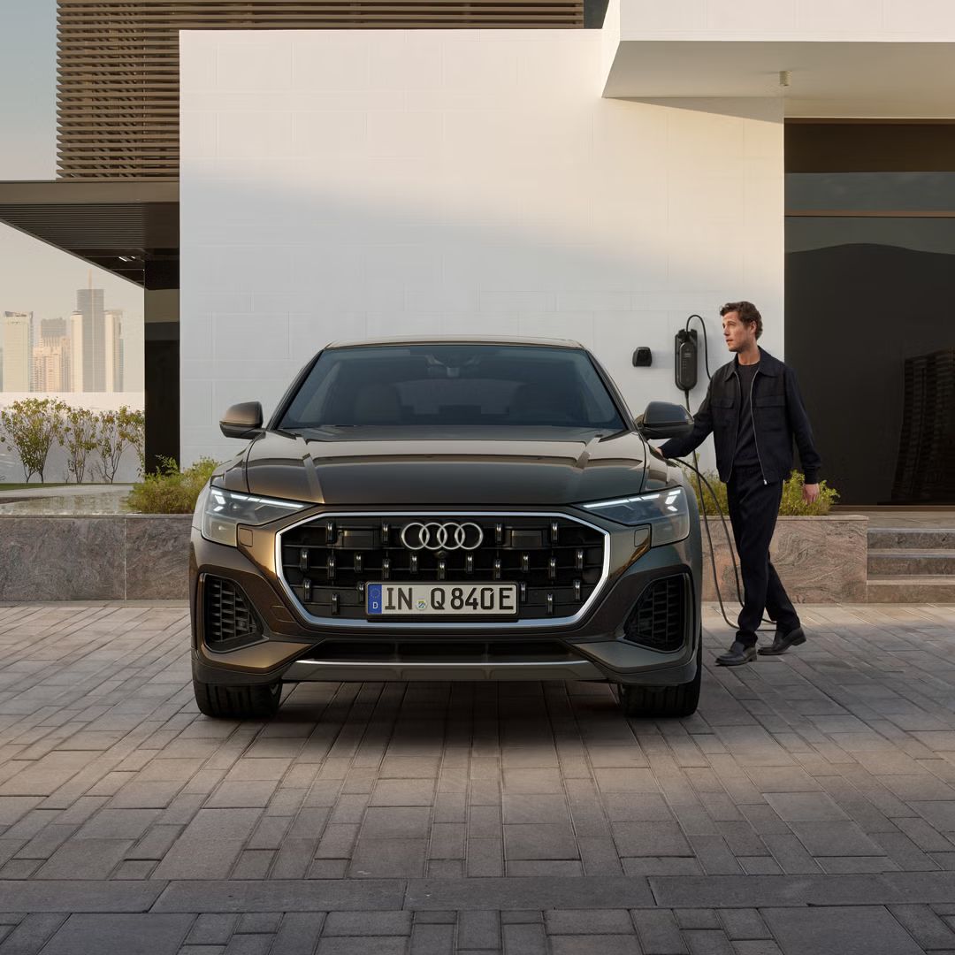 Frontansicht des Q8 SUV e-hybrid, dahinter ein modernes Gebäude. Neben den Fahrzeug steht ein modisch gekleideter junger Mann und hält den Stecker einer am Gebäude angebrachten Ladesäule in der Hand.