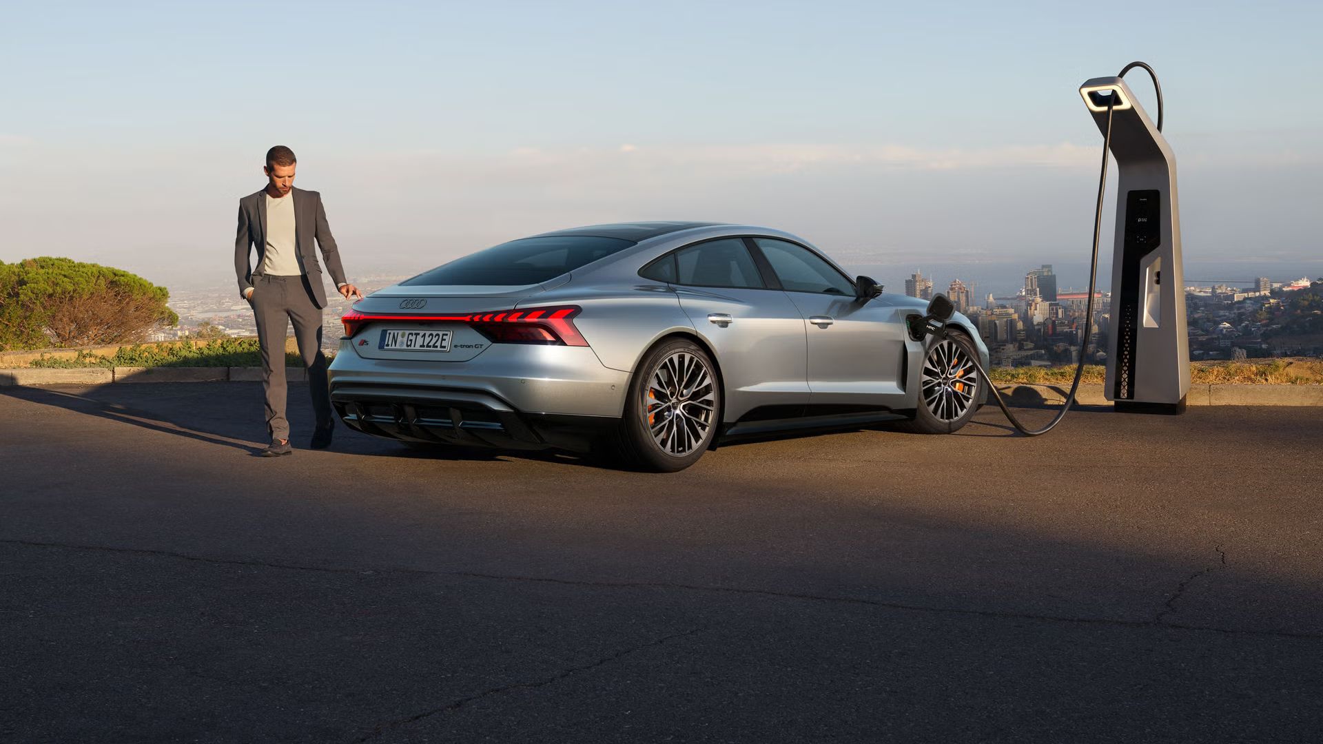 Ein silberner Audi S e-tron GT lädt an einer Ladestation mit Blick auf eine Stadt. Ein Mann im Anzug geht neben dem Auto entlang.