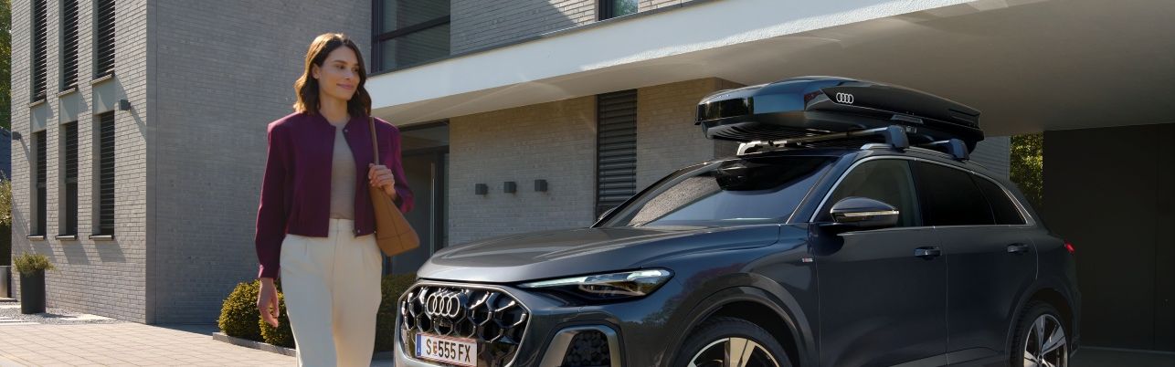 Frau passiert lächelnd einen in der Hauseinfahrt parkenden Audi Q5 mit Dachbox