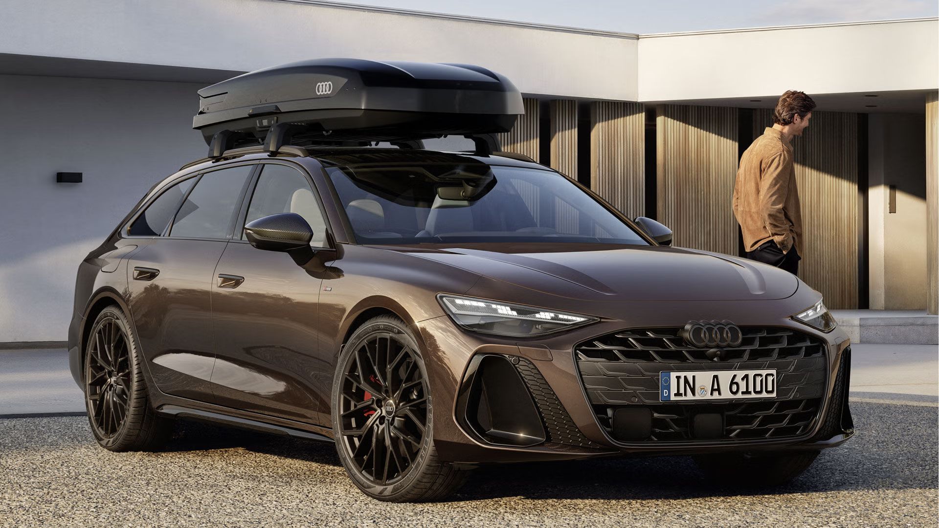 Der A6 Avant steht im schrägen Frontprofil vor einem modernen Haus. Auf dem Dach befindet sich eine Dachbox mit Audi Logo. Links vom Fahrzeug steht ein Mann.