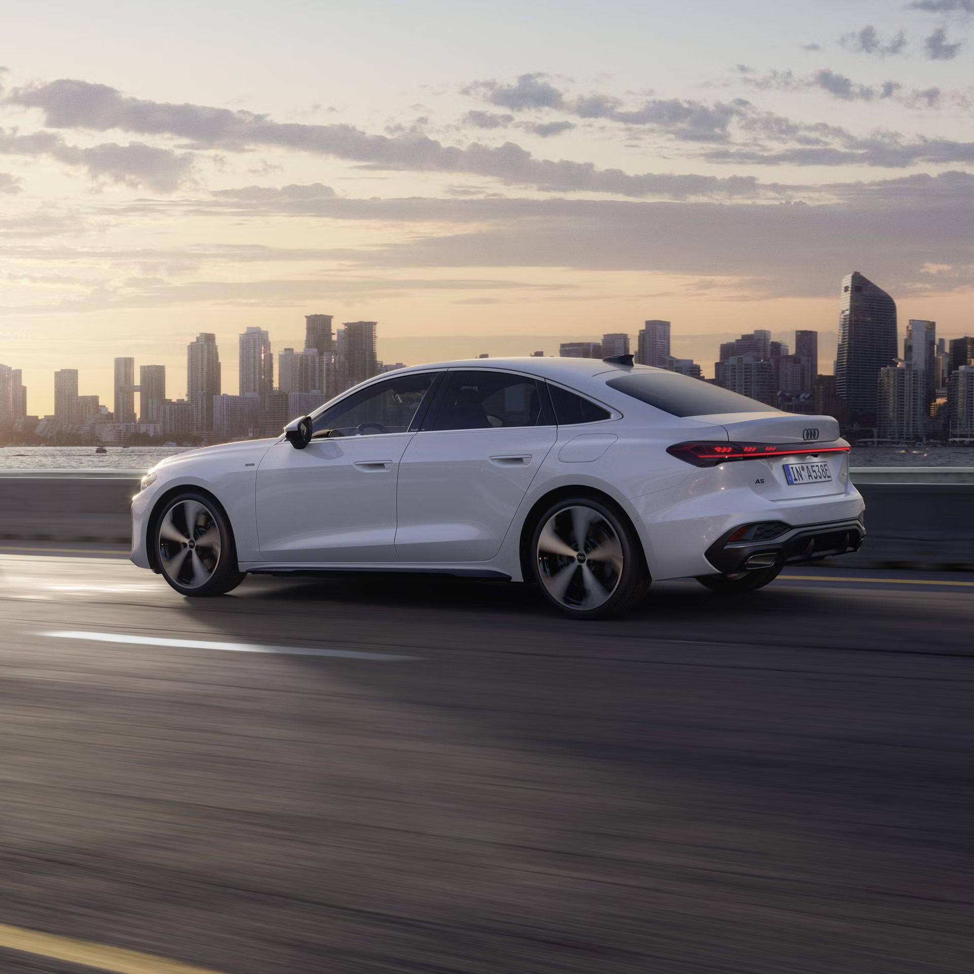 Die A5 Limousine e-hybrid im Seitenprofil, während sie auf einer Straße entlang eines Gewässers fährt. Im Hintergrund ist die Skyline einer Stadt bei Sonnenaufgang zu erkennen.