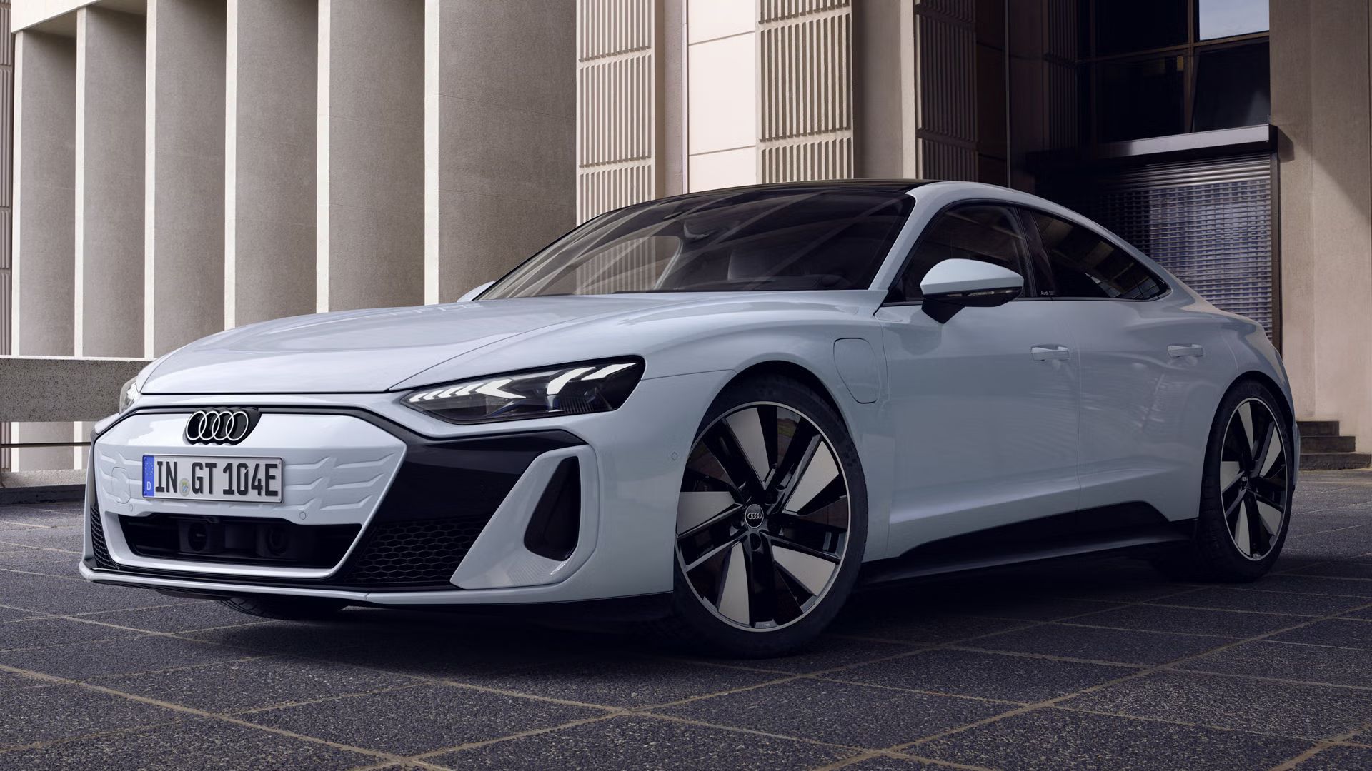 Ein hellblauer Audi e-tron GT quattro schräg von vorn, im Hintergrund ein modernes Gebäude. 