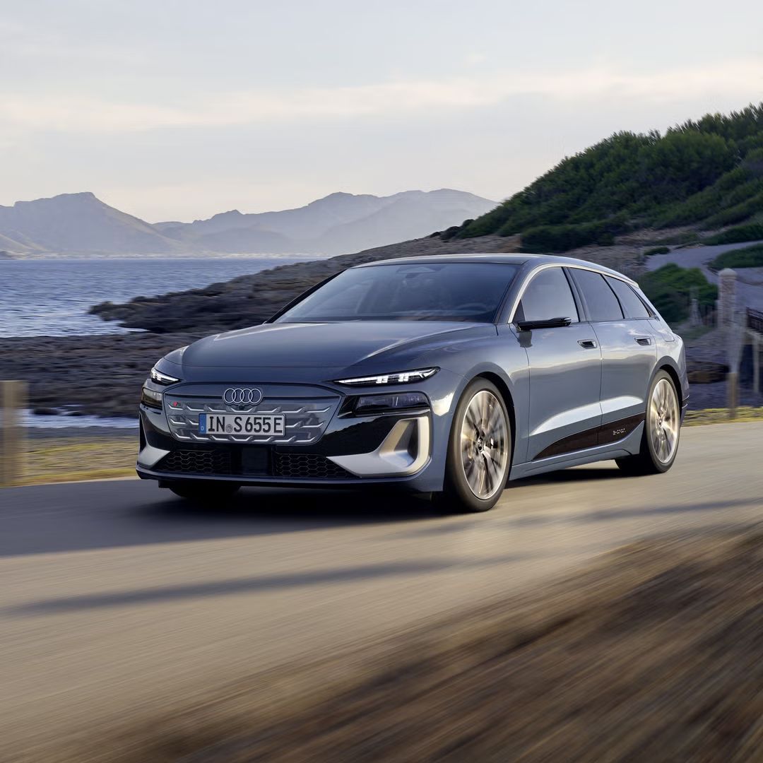 Der S6 Avant e-tron aus schräger Frontansicht, wie er eine Straße an der Küste entlangfährt. Im Hintergrund sind Berge zu sehen. 