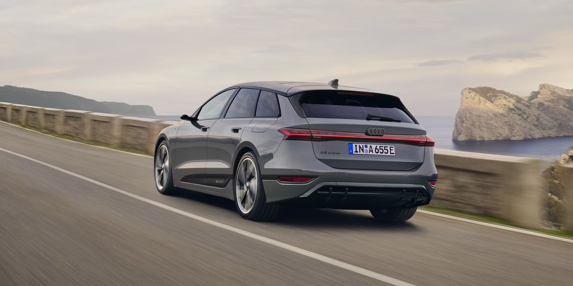 Schräge Heckaufnahme des A6 Avant e-tron, welcher auf einer Straße entlang des Meeres fährt. Im Hintergrund sind Felsen und Berge zu erkennen. 