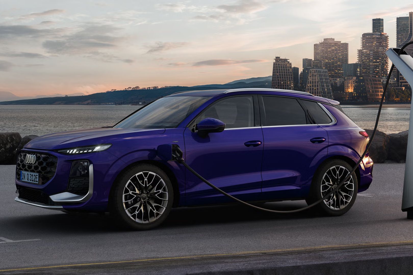 Ein Q3 SUV e-hybrid im Samtviolett Perleffekt an einer Ladestation, im Hintergrund die Skyline einer Küstenstadt.