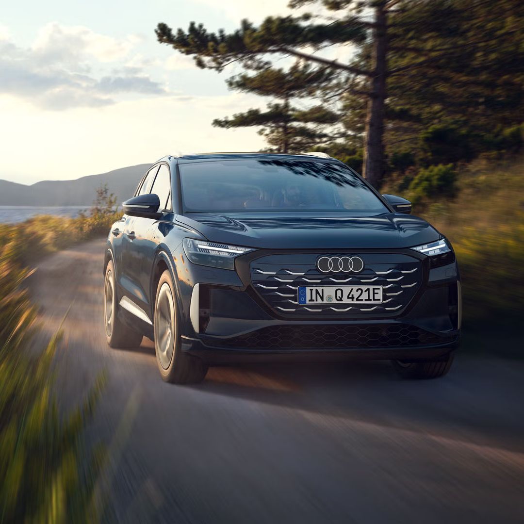 Der Q4 SUV e-tron schräg von vorn in voller Fahrt auf einer Landstraße an einem Waldsee, im Hintergrund eine bergige Landschaft.