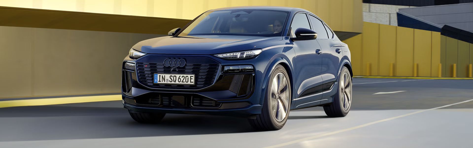 Schräges Frontprofil des SQ6 Sportback e-tron, welcher auf einer Straße durch die Stadt fährt. Im Hintergrund sieht man eine gelbe Gebäudefassade.