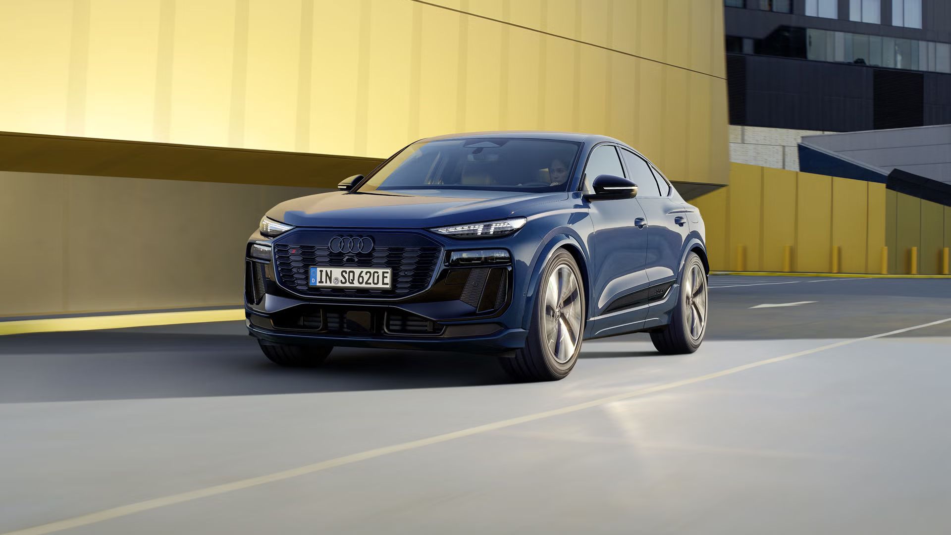 Schräges Frontprofil des SQ6 Sportback e-tron, welcher auf einer Straße durch die Stadt fährt. Im Hintergrund sieht man eine gelbe Gebäudefassade.
