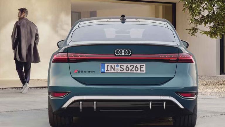 Der S6 Sportback e-tron im Heckprofil. Das Fahrzeug ist dem Eingangsbereich eines modernen Gebäudes zugewandt. Vor dem Auto befindet sich ein junger Mann, der auf das Gebäude zuläuft.