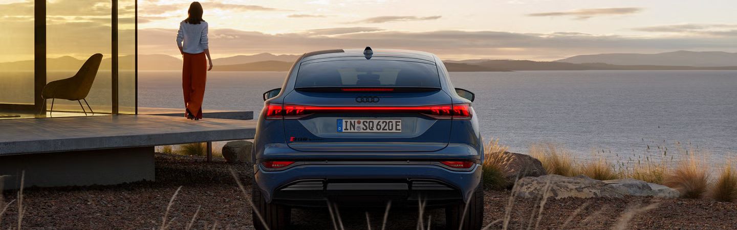 Heckprofil des SQ6 Sportback e-tron, auf Schotter parkend. Im Hintergrund das Meer bei Sonnenuntergang.