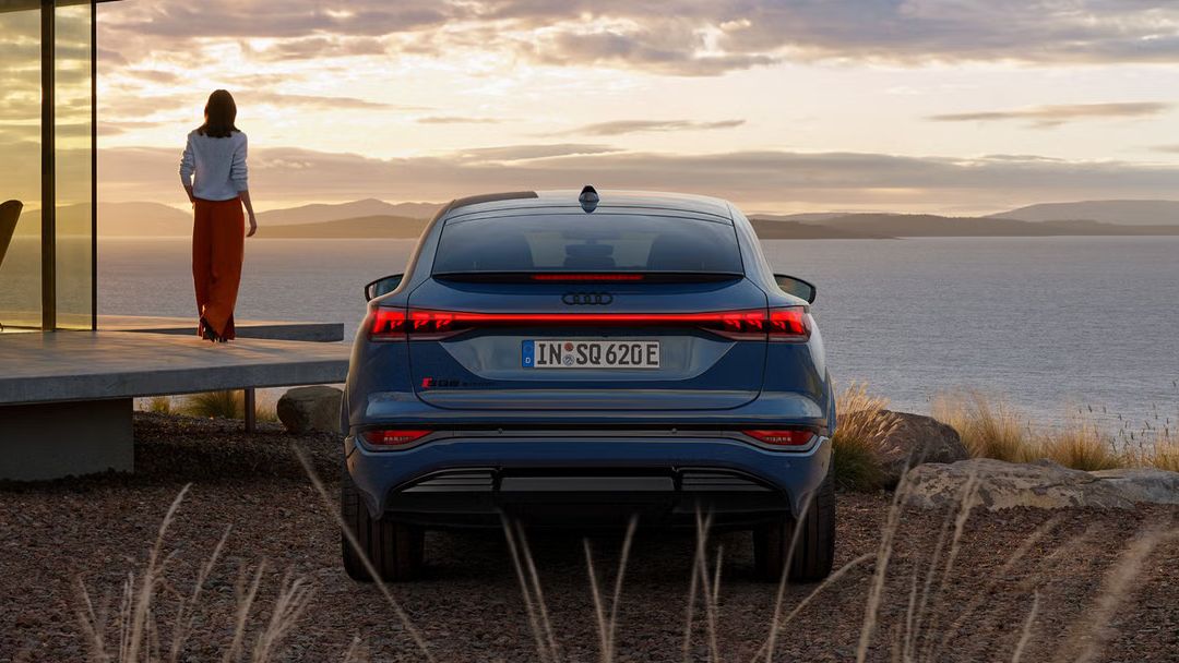 Heckprofil des SQ6 Sportback e-tron, auf Schotter parkend. Im Hintergrund das Meer bei Sonnenuntergang.