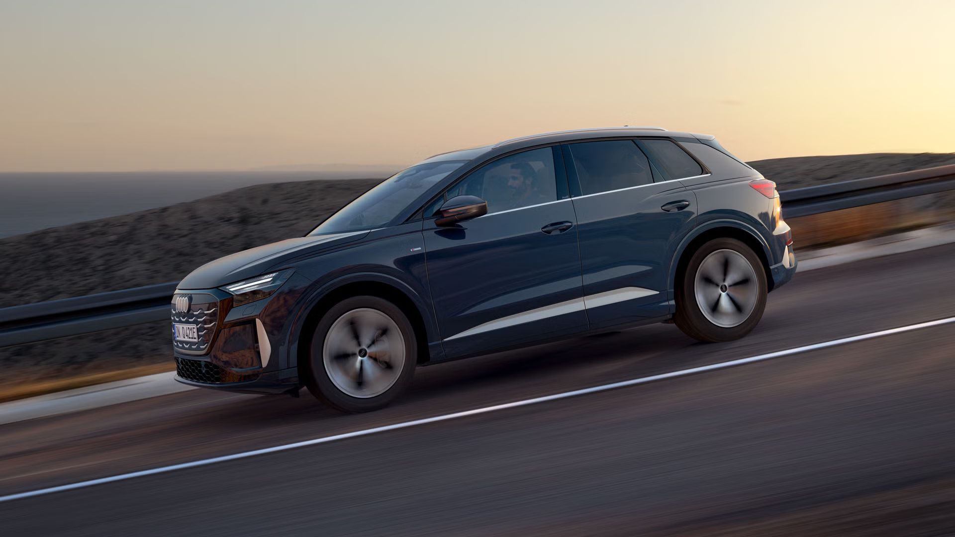 Blick von der Seite auf den Q4 SUV e-tron bei der Fahrt auf abschüssiger Straße im dämmrigen Licht. Im Hintergrund das Meer.
