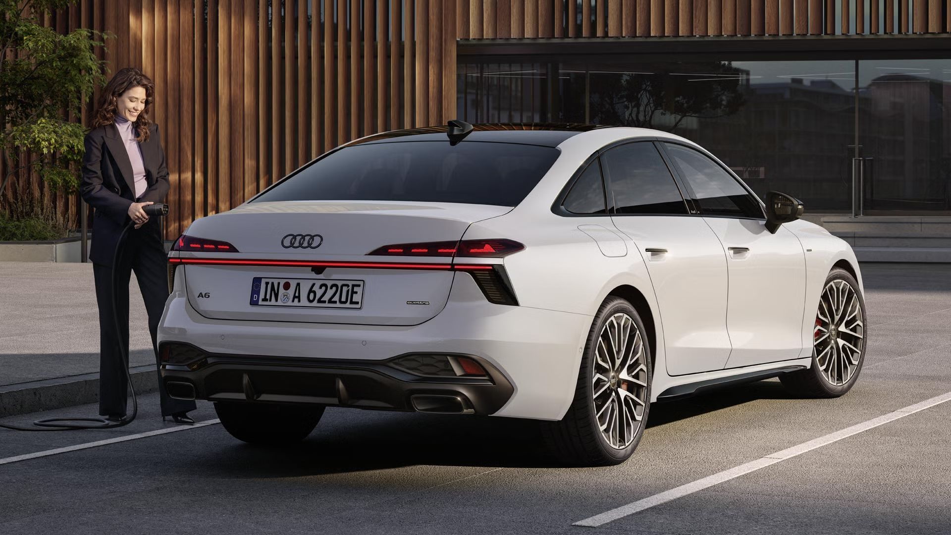 Die A6 Limousine e-hybrid in weiß steht auf einem Parkplatz vor einer Gebäudefassade. Eine junge Frau schließt die A6 Limousine e-hybrid an eine Ladestation an.
