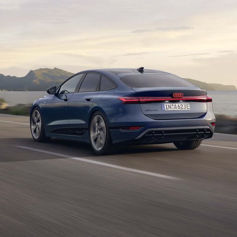 Der A6 Sportback e-tron aus schräger Heckansicht fährt eine Straße parallel zum Ufer entlang. Im Hintergrund sind Berge zu erkennen. 