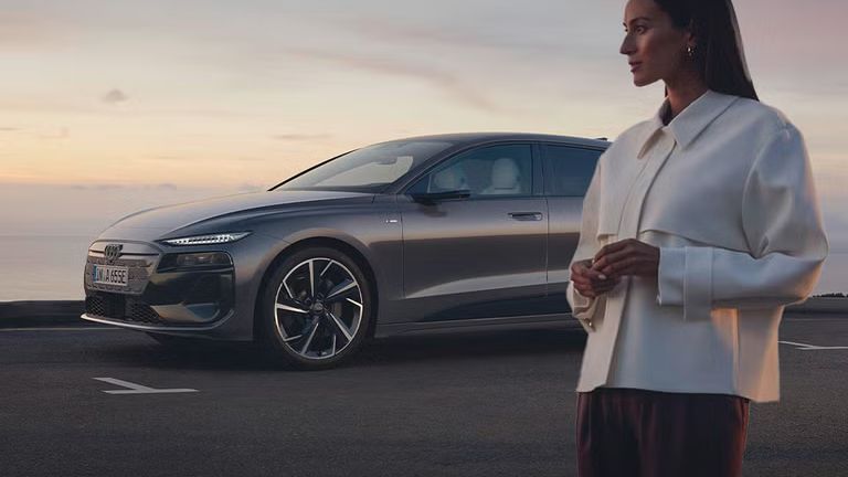Grauer Audi A6 e-tron parkt vor Meer bei Sonnenuntergang, Frau in weißer Jacke steht daneben