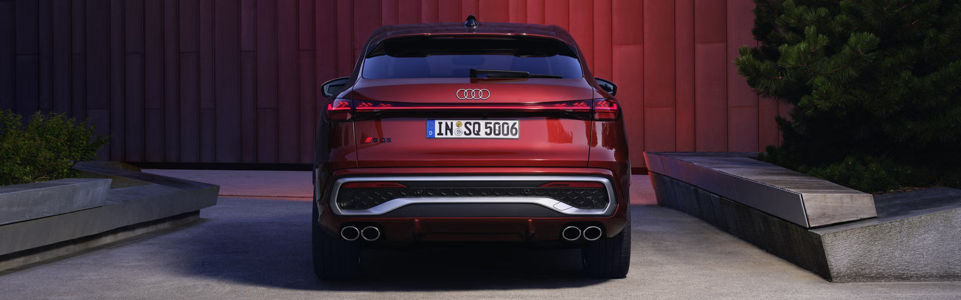 Heckprofil des roten SQ5 Sportback, welcher in einem Stadtpark vor einer Gebäudefassade steht und diese rot anleuchtet.