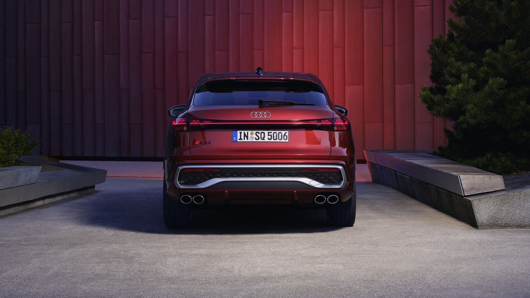 Heckprofil des roten SQ5 Sportback, welcher in einem Stadtpark vor einer Gebäudefassade steht und diese rot anleuchtet.