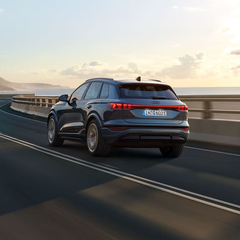 Schräge Heckaufnahme des Q6 SUV e-tron, welche über eine kurvige Straße am Meer fährt. Im Hintergrund sind Berge zu sehen.