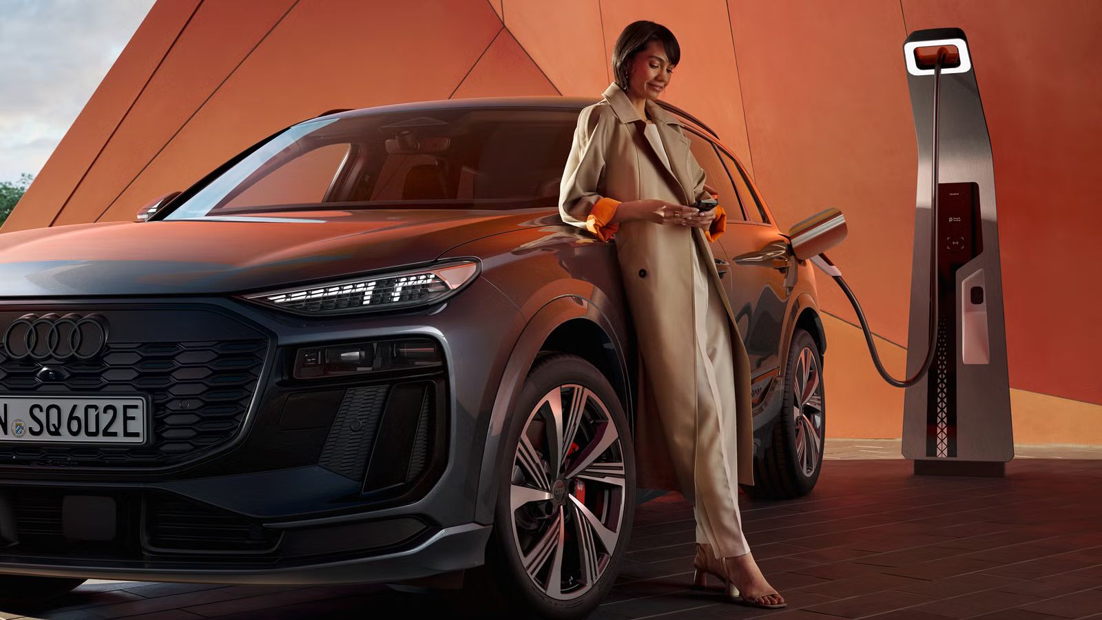 Der SQ6 SUV e-tron steht vor einer orangenen Gebäudefassade und ist dabei an eine Ladestation angeschlossen. An der Seite des Fahrzeugs lehnt sich eine junge Frau an und schaut auf ihr Handy. 
