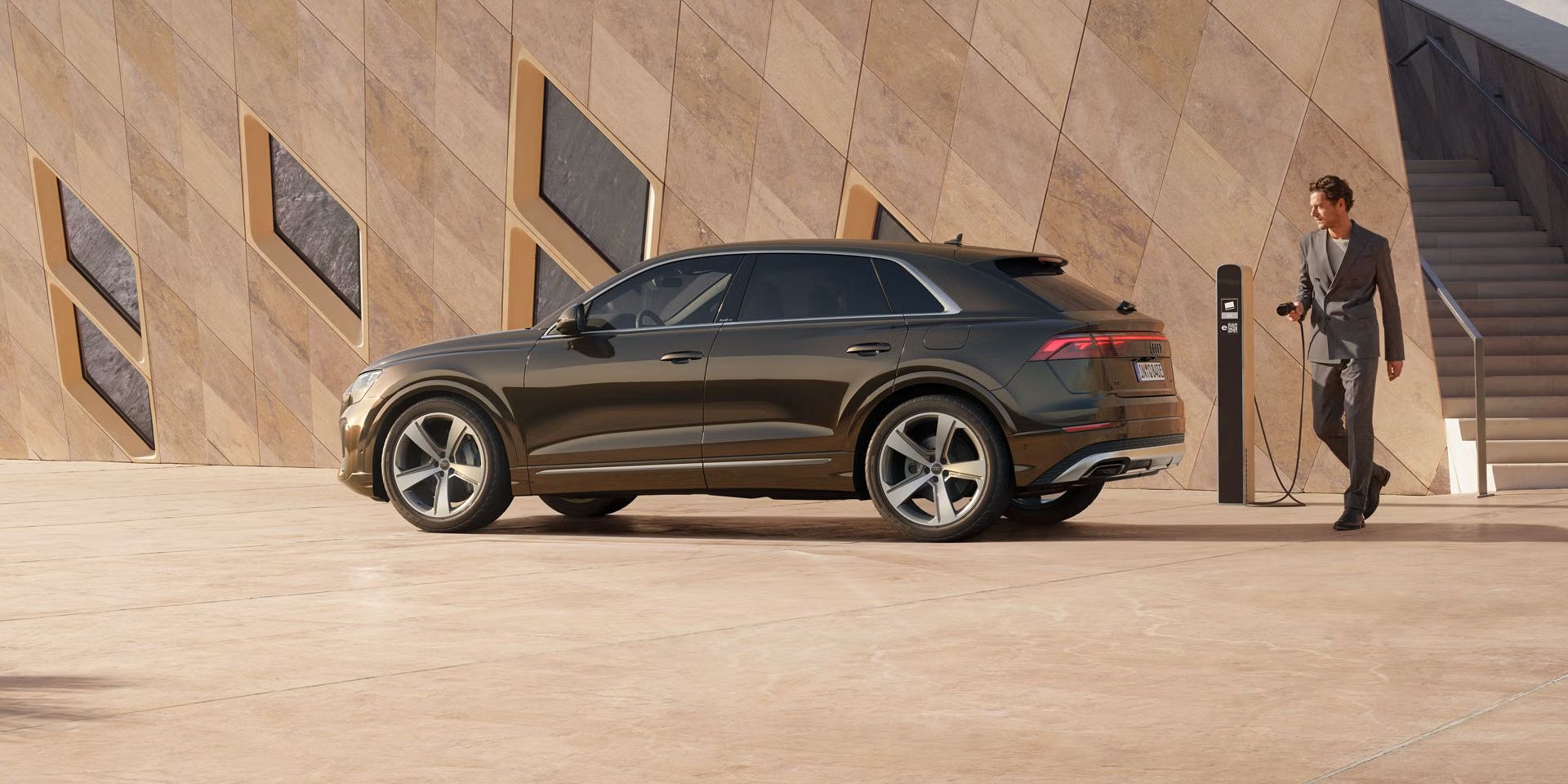 Der Q8 SUV e-hybrid schräg von der Seite parkt vor einem futuristischen Gebäude. Ein modisch gekleideter junger Mann steht hinter dem Fahrzeug und hält den Stecker einer Ladesäule in der Hand.