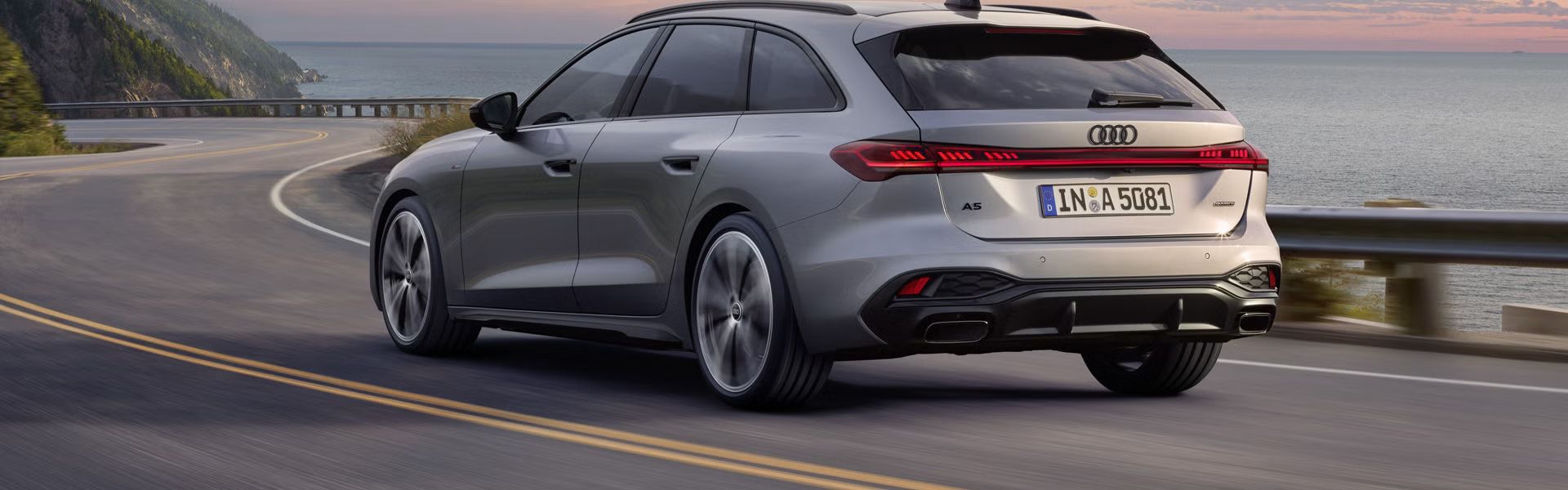 Der A5 Avant in einer schrägen Heckperspektive, während er bei Sonnenuntergang eine kurvige Küstenstraße entlangfährt.