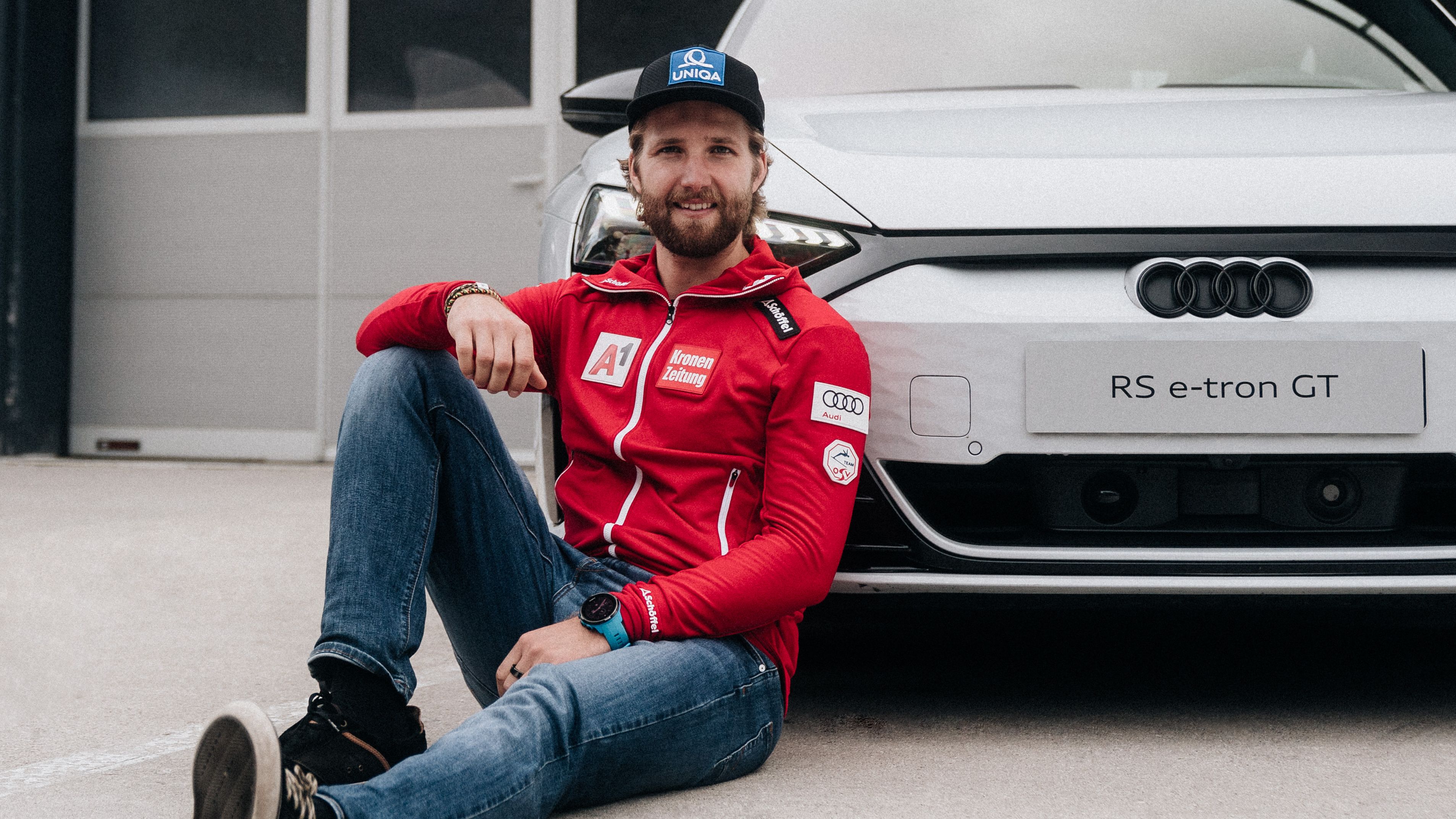 Skirennläufer Marco Schwarz sitzt vor einem weißen Audi RS e-tron GT. 