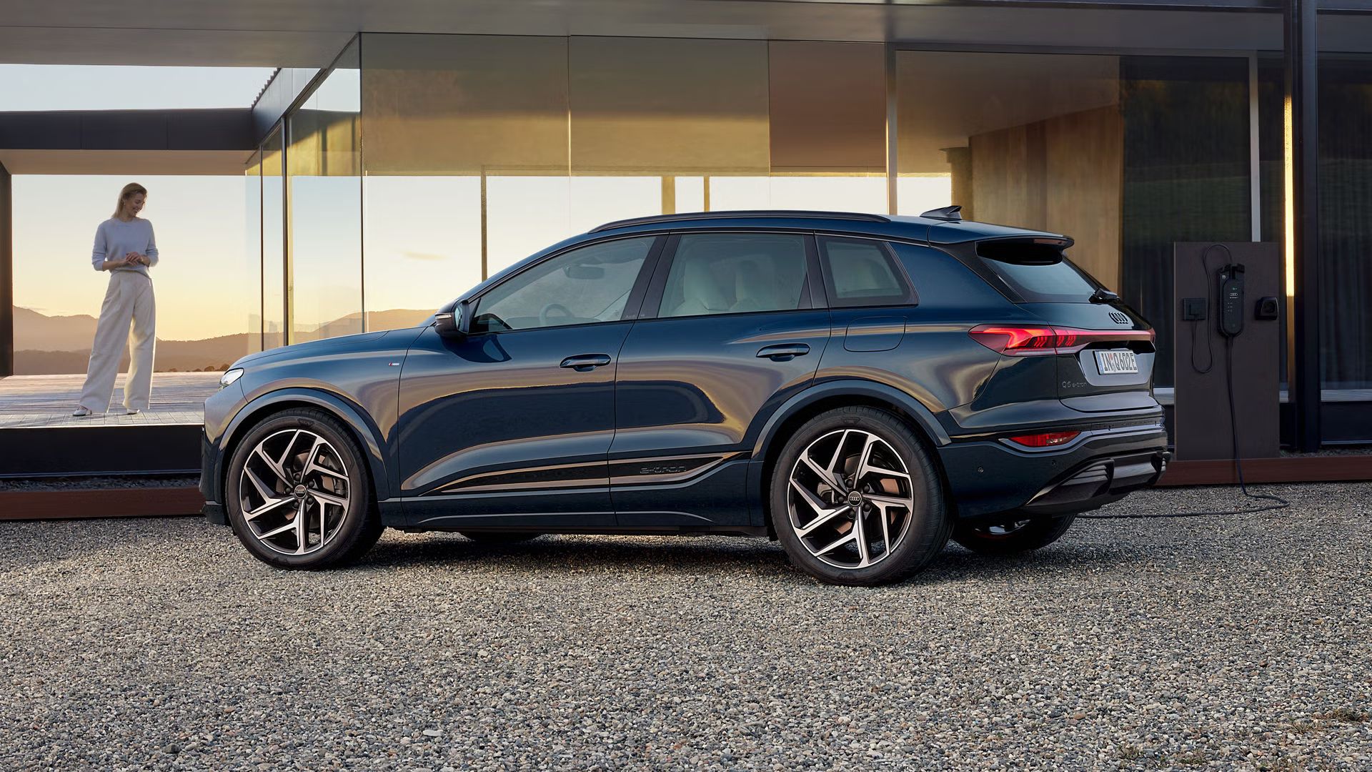 Der Q6 SUV e-tron steht seitlich auf der Kiesfläche vor einem modernen Haus und ist an eine Ladestation angeschlossen. Neben dem Haus steht eine junge Frau.