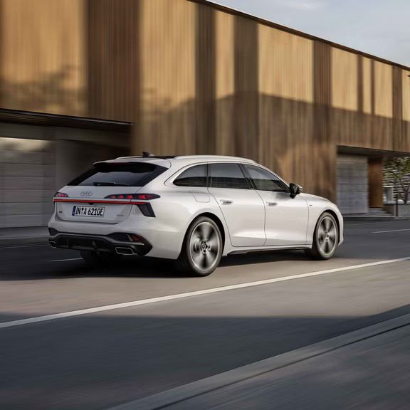 Blick von schräg hinten auf den A6 Avant e-hybrid, der auf einer Straße vor einem Gebäude entlangfährt.