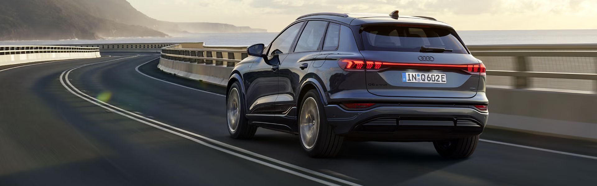 Schräge Heckaufnahme des Q6 SUV e-tron, welche über eine kurvige Straße am Meer fährt. Im Hintergrund sind Berge zu sehen.