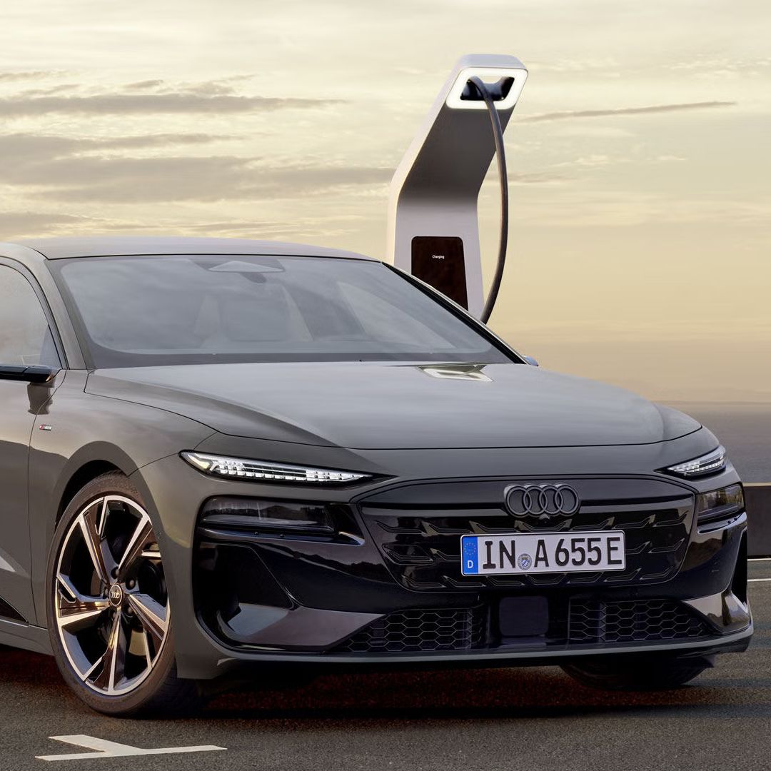 Der A6 Avant e-tron steht rückwärts auf einem Parkplatz und ist dabei an eine Ladestation angeschlossen. Im Hintergrund sind das Meer, sowie Berge zu erkennen. 