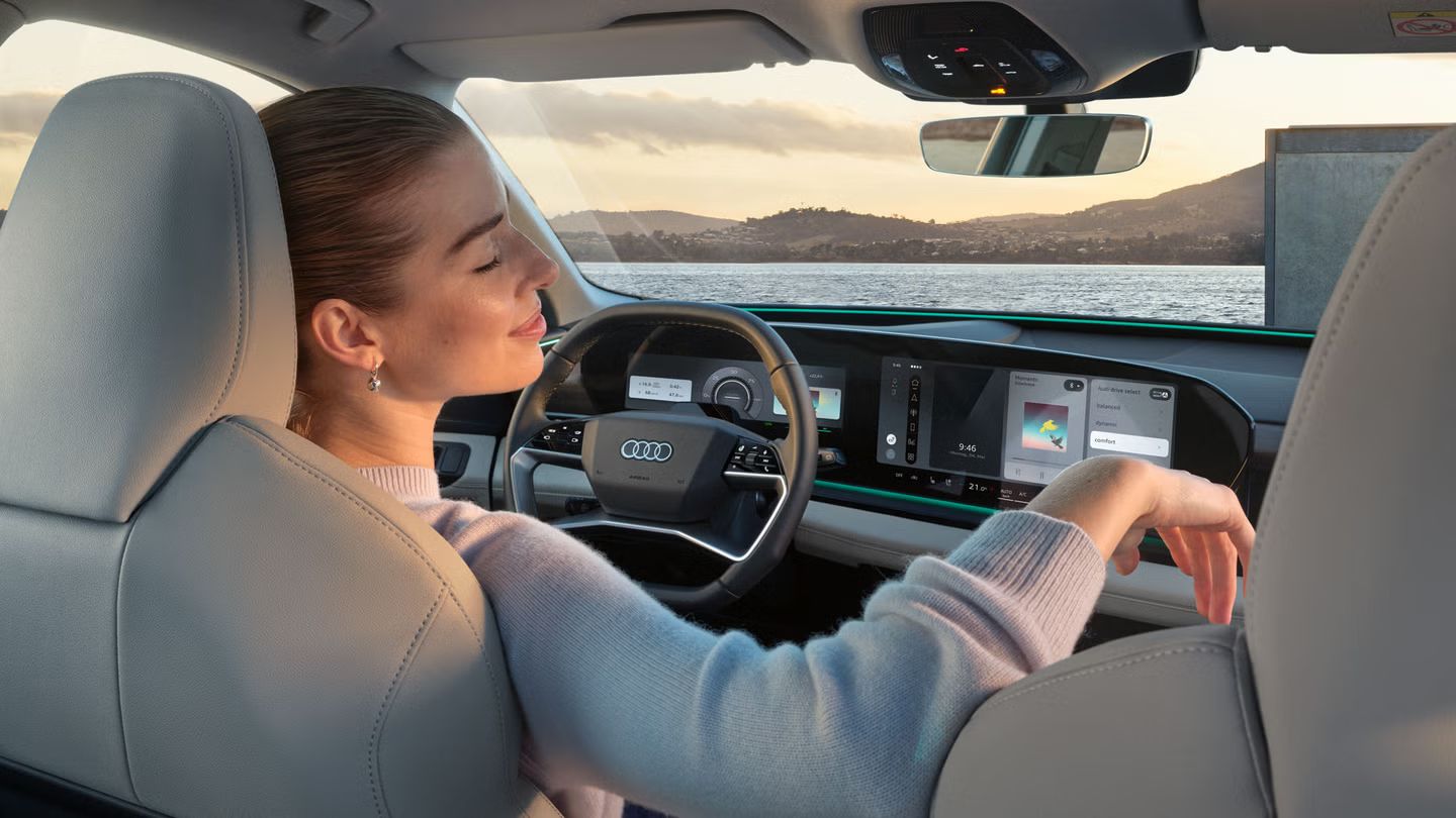 Innenraum des Q6 SUV e-tron aus Rücksitzperspektive. Eine junge Frau sitzt auf dem Fahrersitz und lehnt sich mit geschlossenen Augen zurück. Durch die Windschutzscheibe sieht man das Meer und Berge.