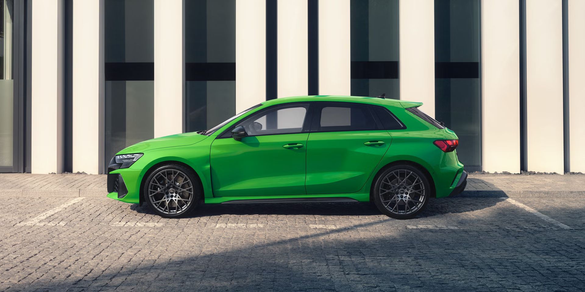 Der RS 3 Sportback von der Seite parkend auf Kopfsteinpflaster, im Hintergrund Umrisse eines modernen Gebäudes.