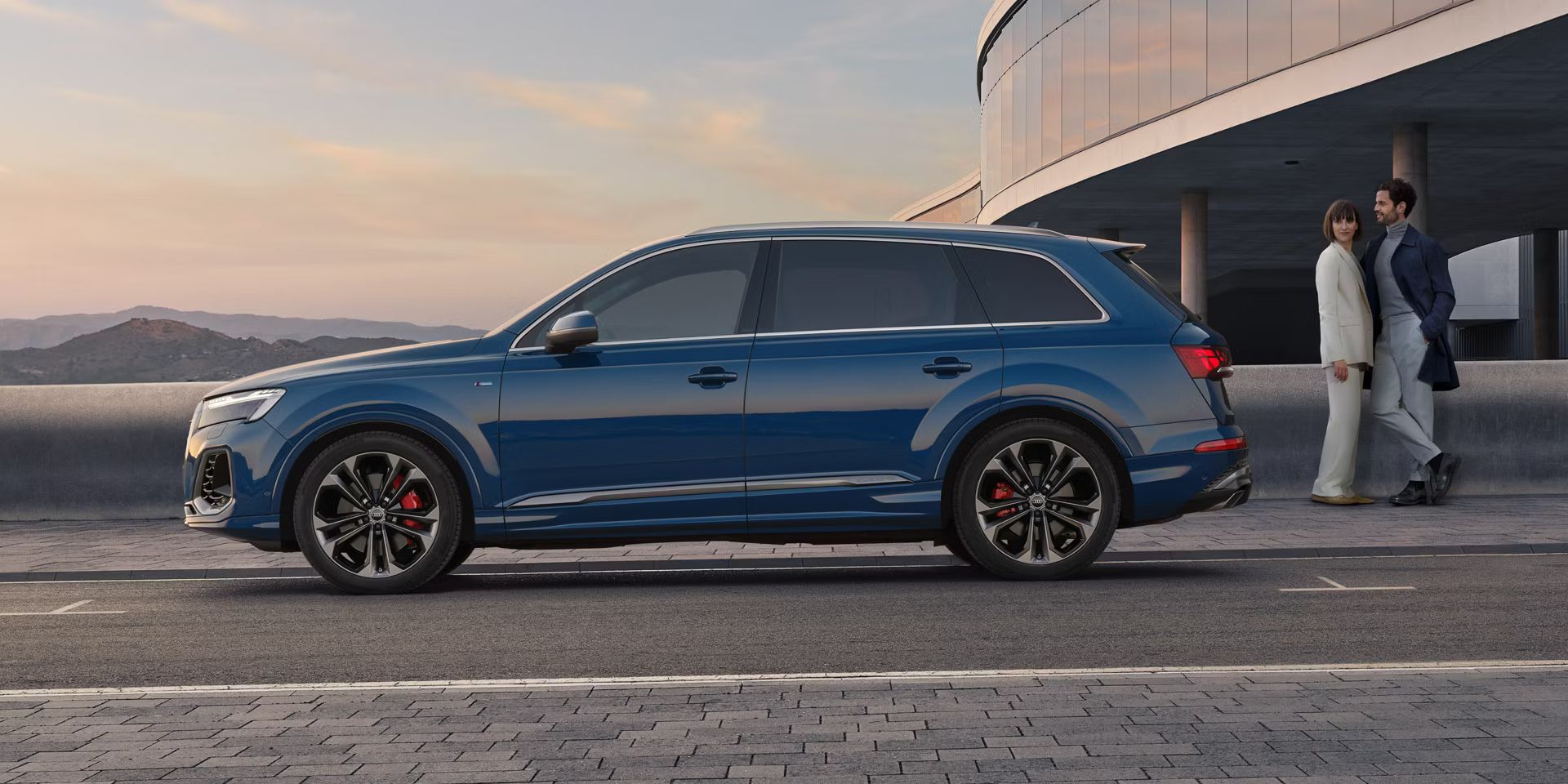 Seitenansicht eines blauen Audi Q7, eine Frau und ein Mann stehen hinter dem Fahrzeug.