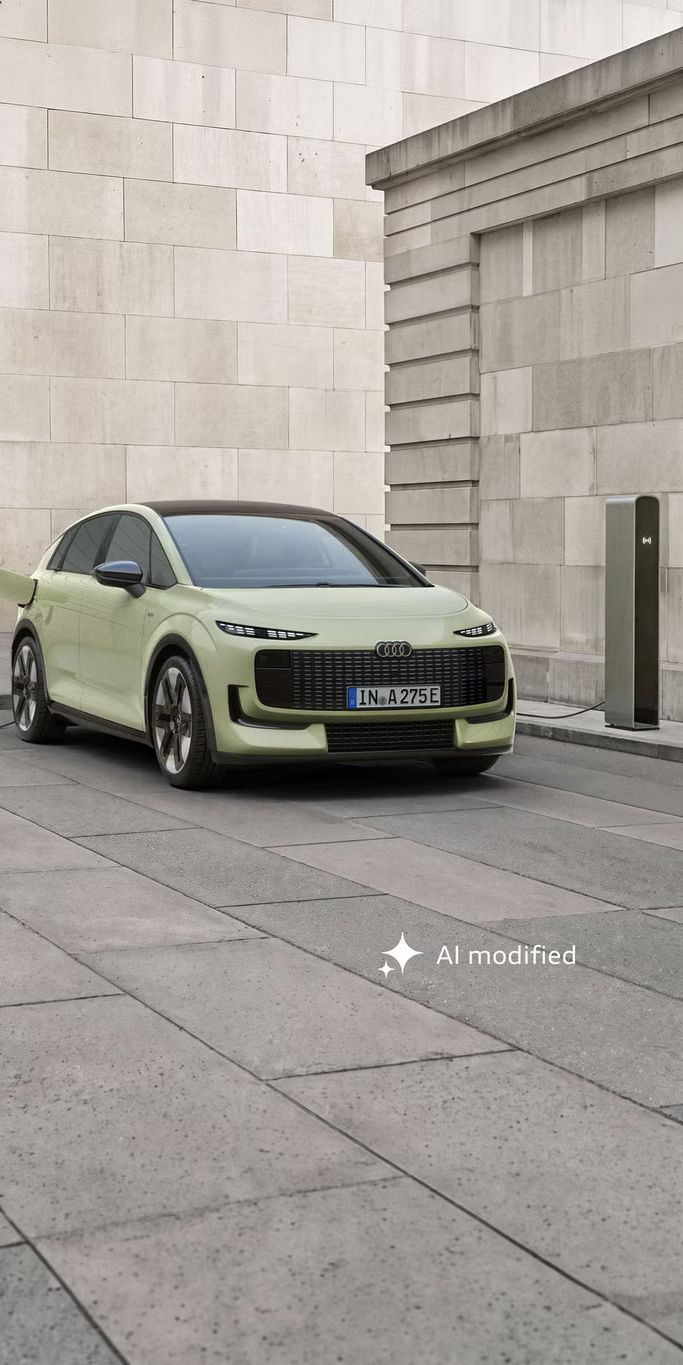 Der Audi A2 e-tron, frontal von hinten fotografiert, parkt vor einem modernen Gebäude. Das Fahrzeug ist an eine Ladesäule angeschlossen.