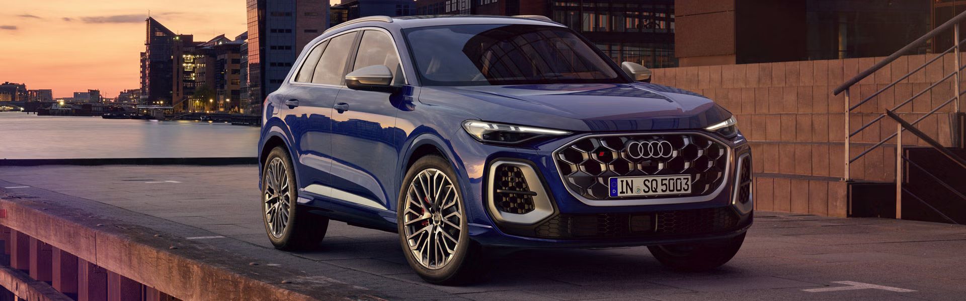 Der SQ5 SUV steht vor der Skyline einer Küstenstadt. Davor befindet sich eine junge, modische Frau. 