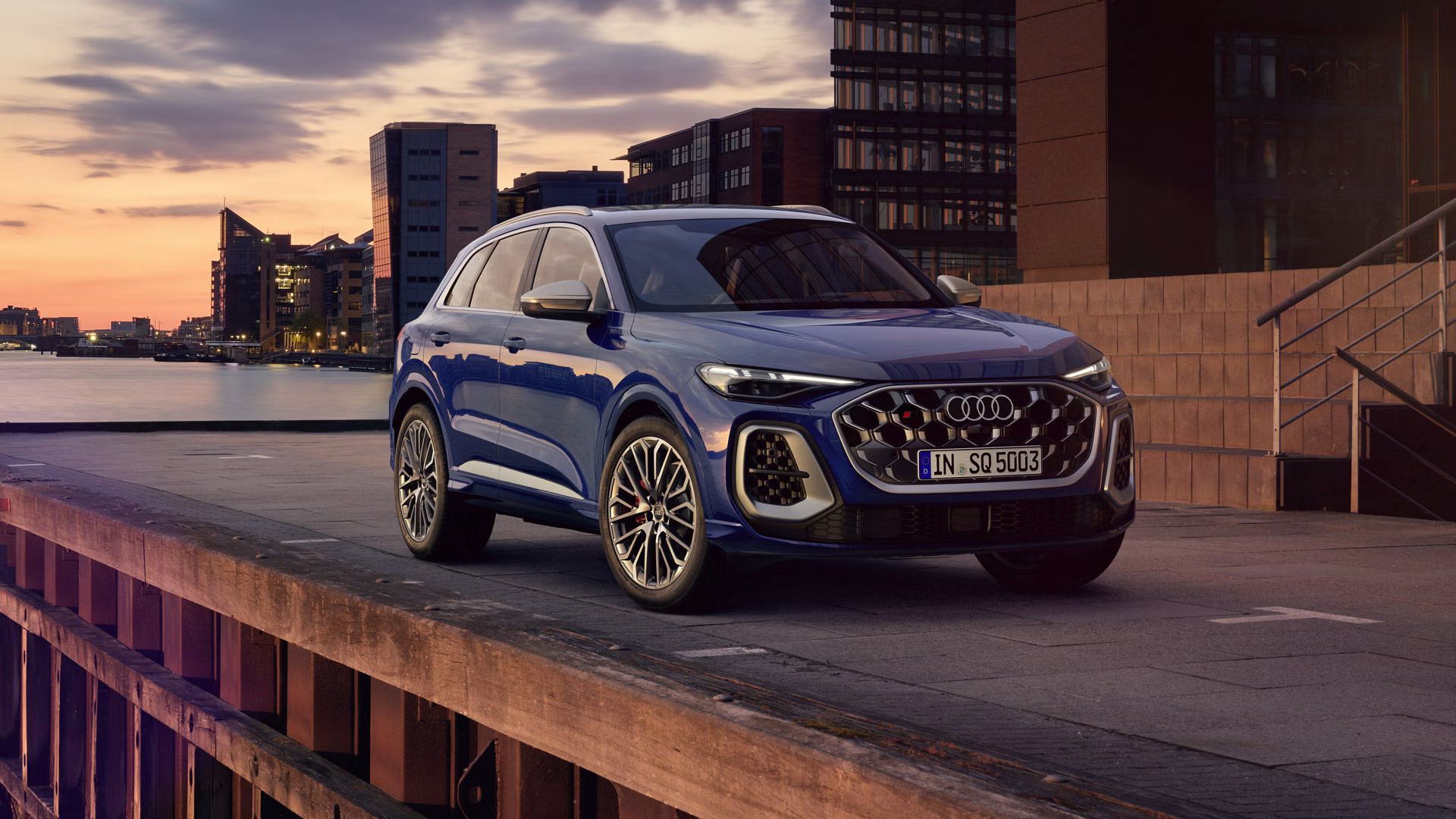 Der SQ5 SUV steht vor der Skyline einer Küstenstadt. Davor befindet sich eine junge, modische Frau. 