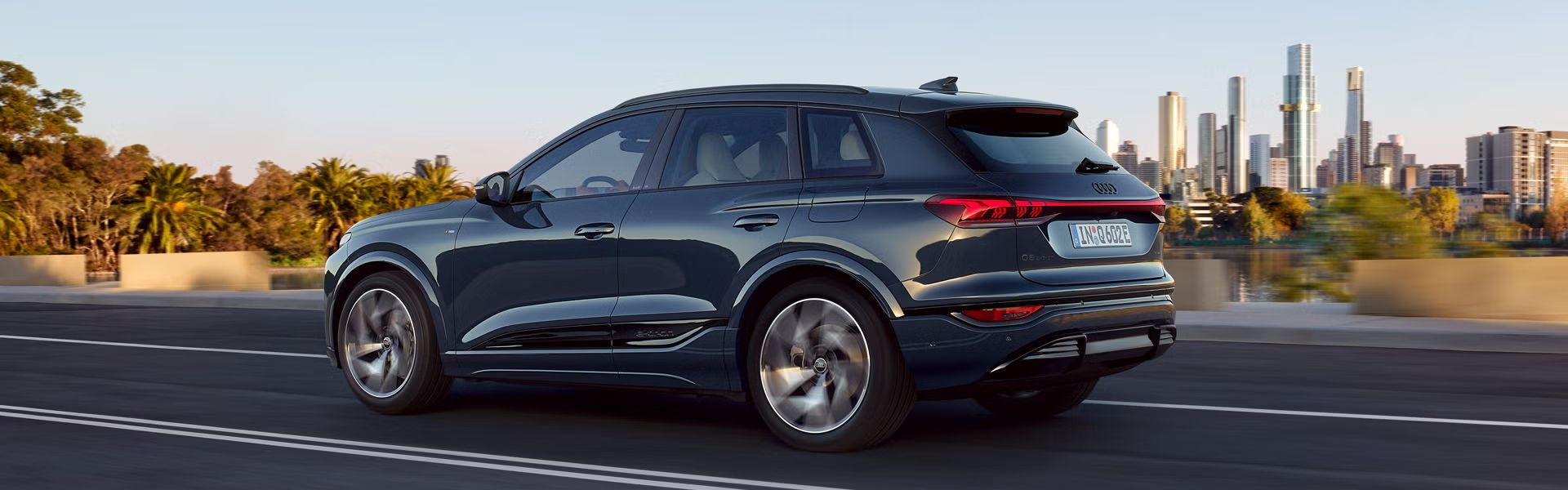 Seitenprofil des Q6 SUV e-tron, welcher auf einer Straße an einem Fluss fährt. Im Hintergrund ist die Skyline einer Großstadt zu sehen.