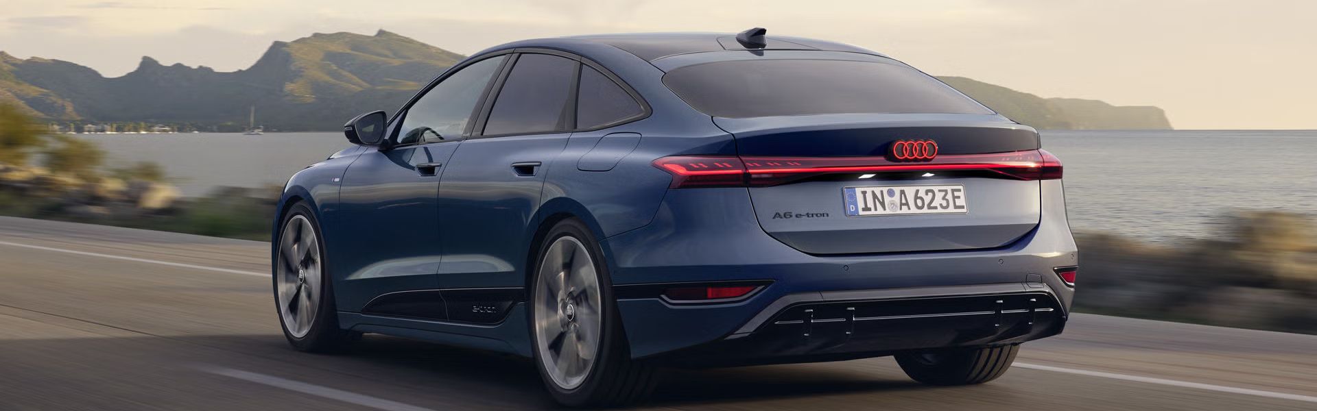 Der A6 Sportback e-tron aus schräger Heckansicht fährt eine Straße parallel zum Ufer entlang. Im Hintergrund sind Berge zu erkennen. 