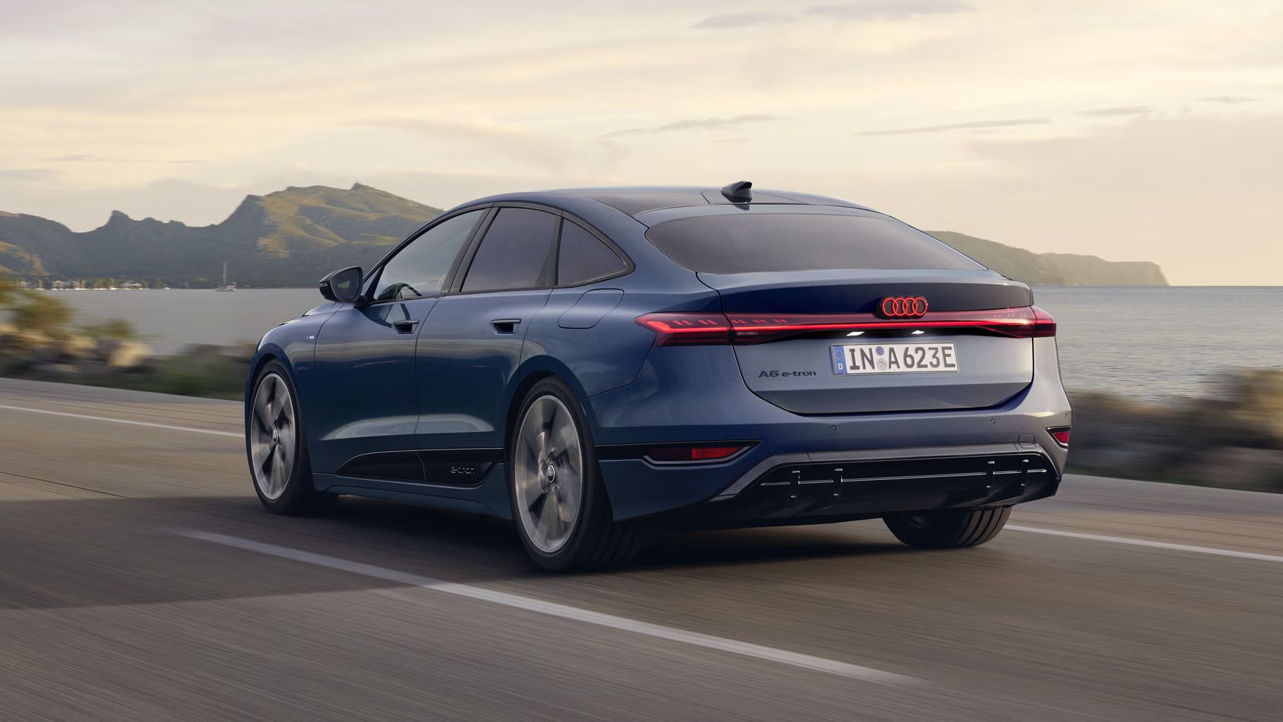 Der A6 Sportback e-tron aus schräger Heckansicht fährt eine Straße parallel zum Ufer entlang. Im Hintergrund sind Berge zu erkennen. 