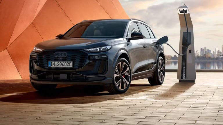 Seitliche Front des SQ6 SUV e-tron, welcher neben einer orangenen Gebäudefassade an eine Ladestation angeschlossen ist. Im Hintergrund ist das Meer, sowie die Skyline einer Stadt zu erkennen.