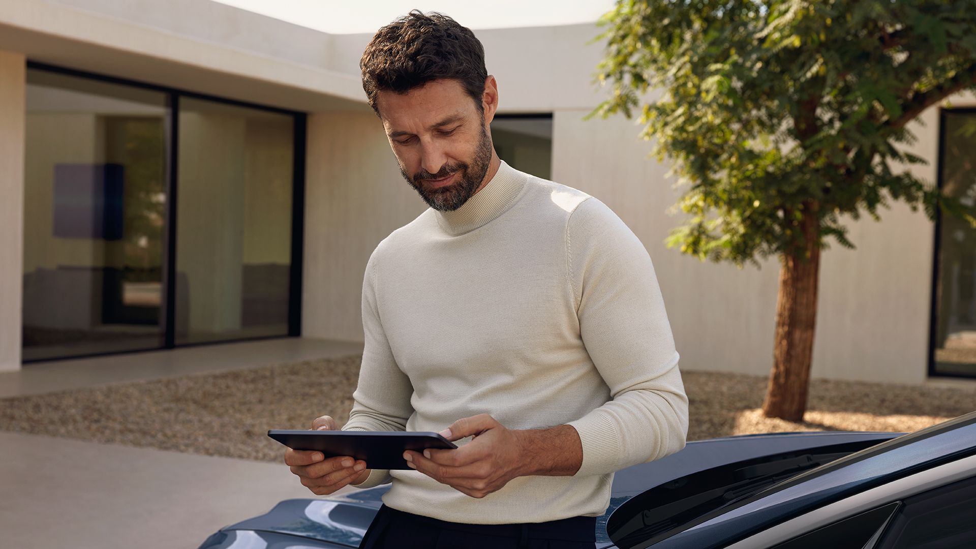 Ein Mann steht im Freien und blickt auf ein Tablet in seinen Händen. Er trägt einen hellen Rollkragenpullover und lehnt sich an einen Audi. Im Hintergrund stehen ein modernes Gebäude mit großen Glasfronten und ein Baum.