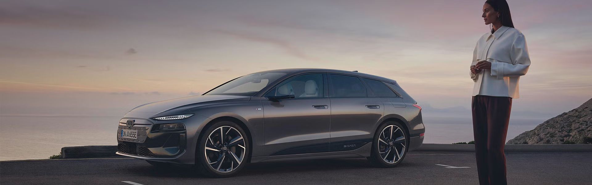 Grauer Audi A6 e-tron parkt vor Meer bei Sonnenuntergang, Frau in weißer Jacke steht daneben