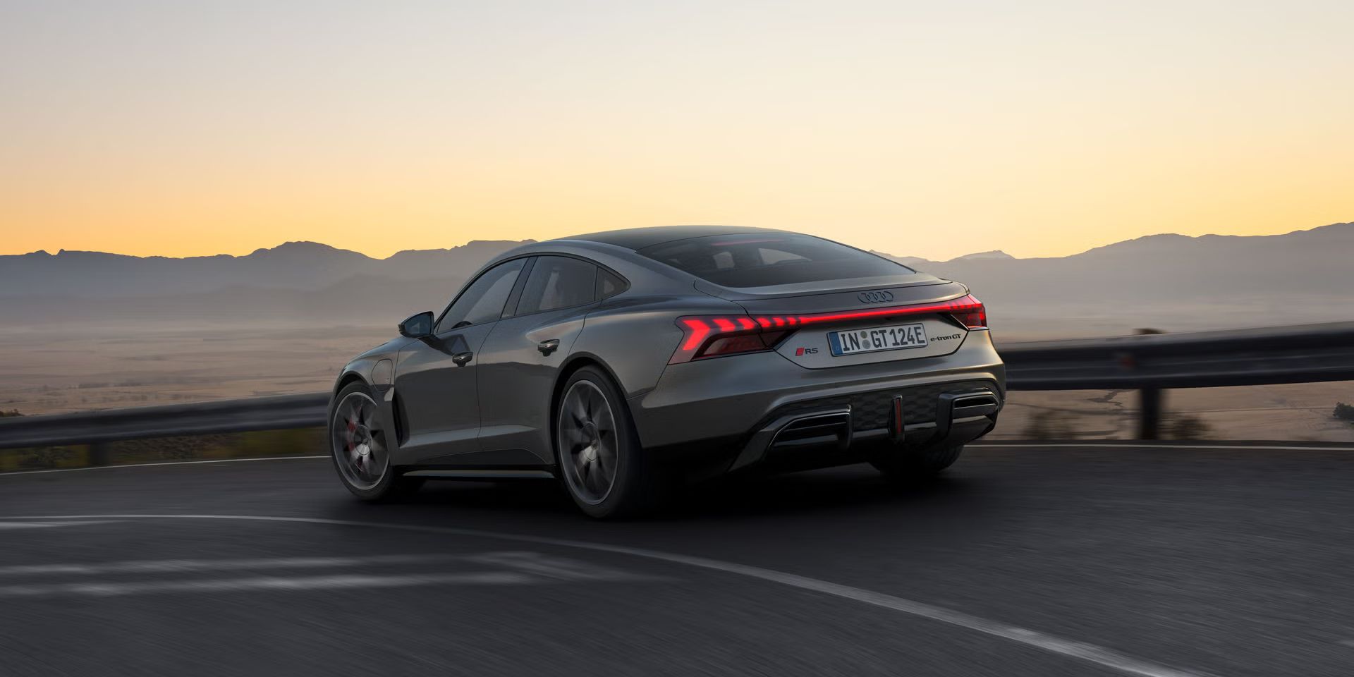 Ein grauer Audi RS e-tron GT fährt auf einer kurvigen Bergstraße bei Sonnenuntergang, mit leuchtenden Rücklichtern.