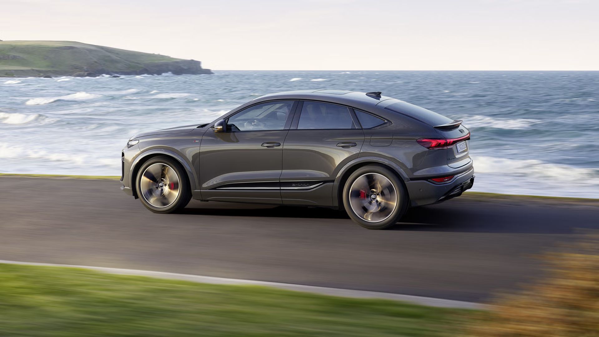 Seitliche Aufnahme des Q6 Sportback e-tron, welcher auf einer Straße entlang des Meeres fährt.