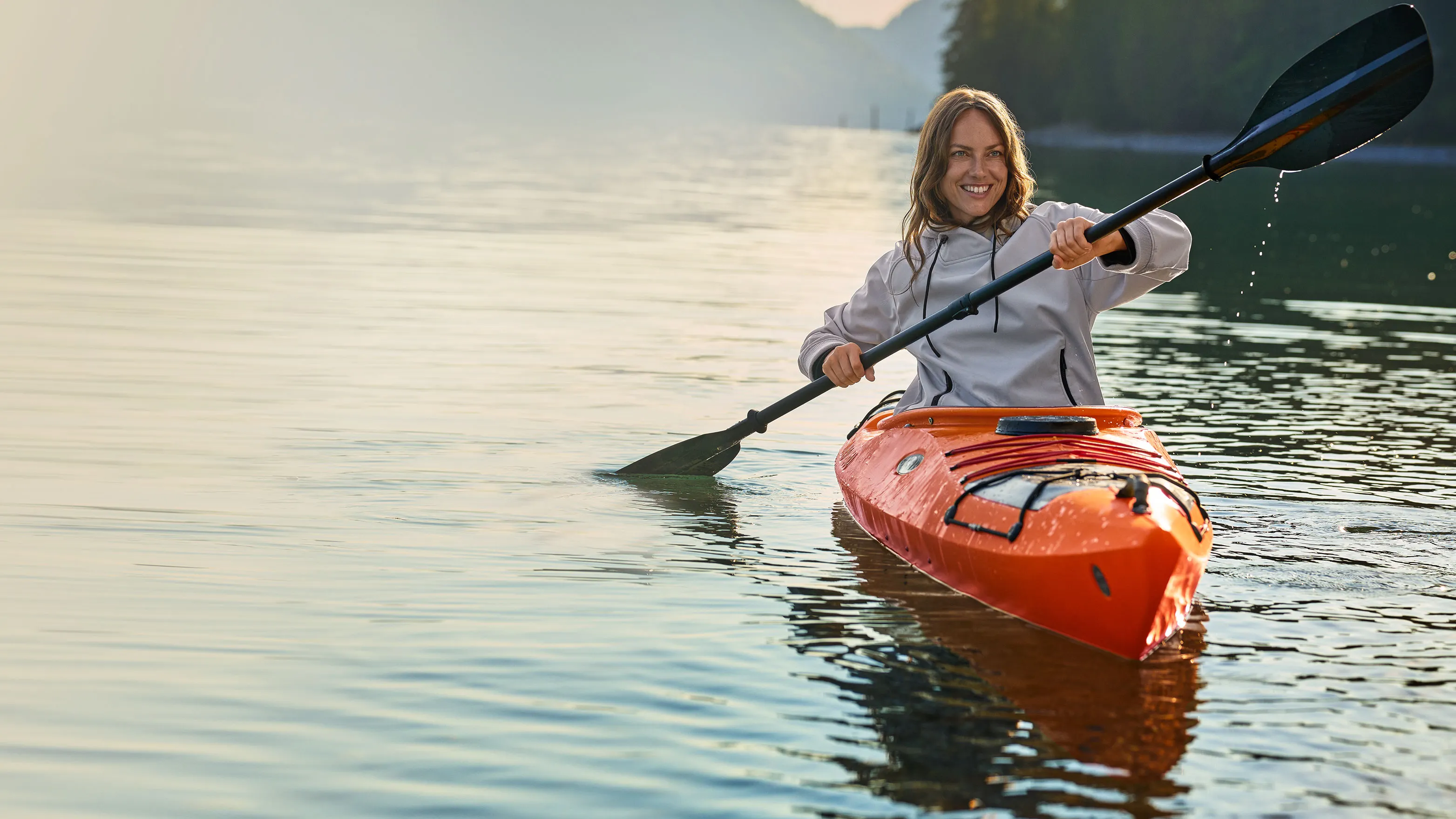 Eine Person in einem orangefarbenen Kajak paddelt auf einem ruhigen See, umgeben von bewaldeten Hügeln und Bergen in der Ferne.