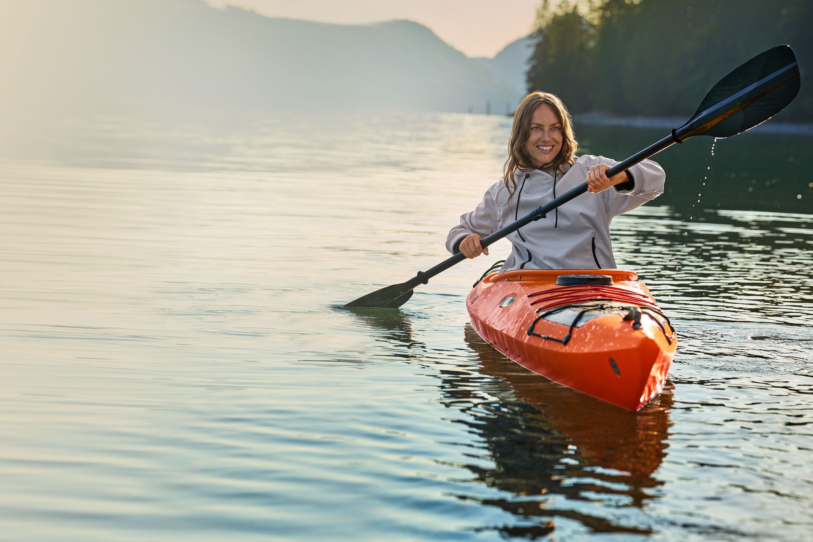Eine Person in einem orangefarbenen Kajak paddelt auf einem ruhigen See, umgeben von bewaldeten Hügeln und Bergen in der Ferne.