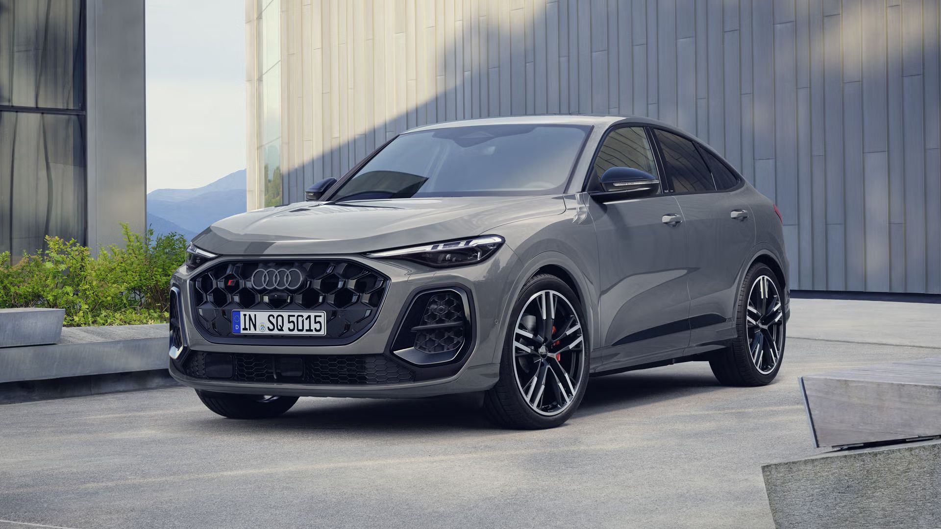Der SQ5 Sportback steht im schrägen Frontprofil vor einem großen Gebäude. Das Fahrzeug ist Nardograu lackiert.